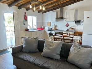 Een gezellige woonkamer met een bank en kussens, een keuken met een wasmachine, en een eethoek met een tafel en stoelen.