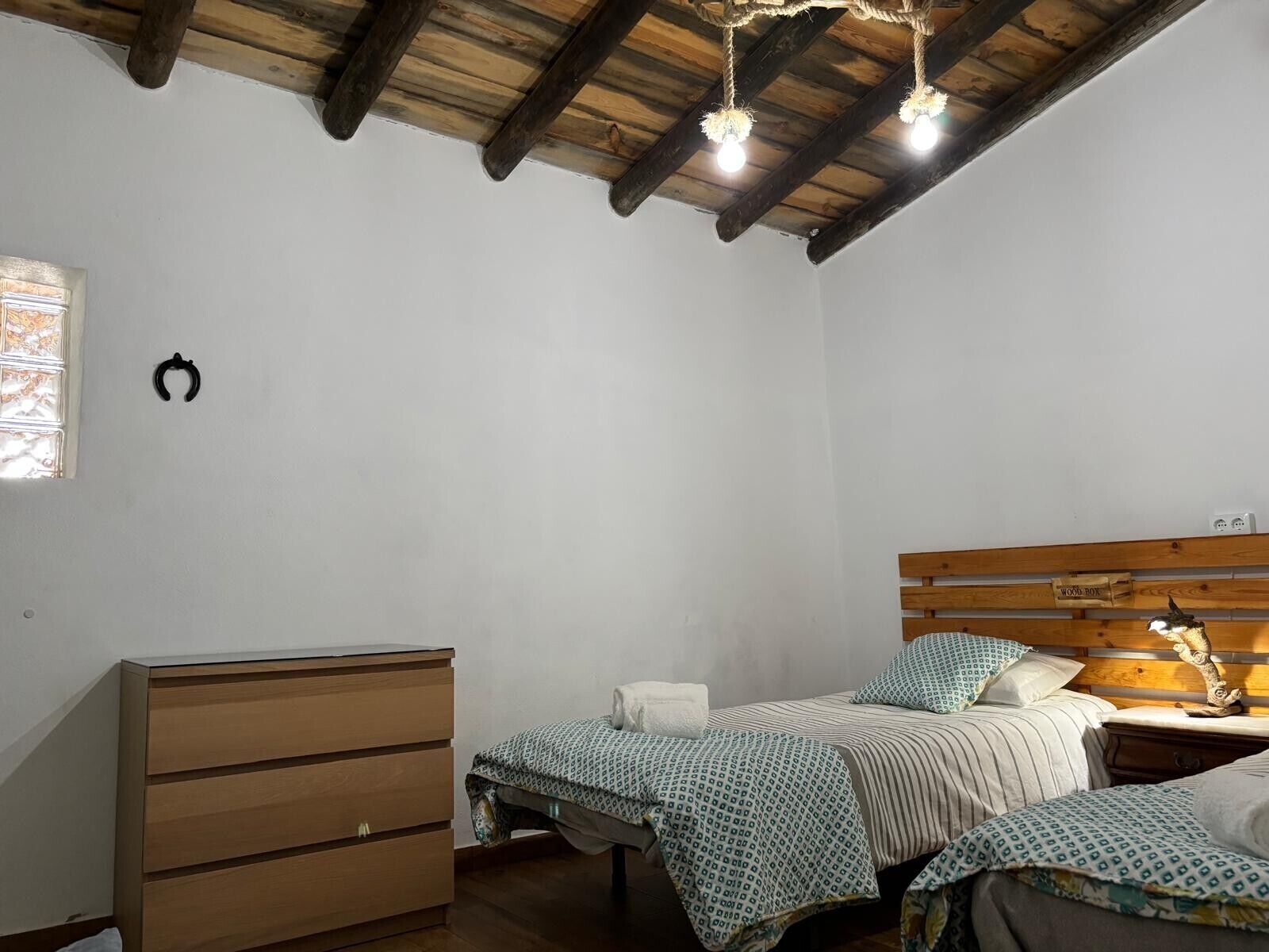 A bedroom with a wooden headboard, two beds with patterned bedding, a wooden dresser, and a hanging light fixture.