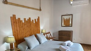 Quarto com cabeceira de madeira, cama com colcha acolchoada, duas luminárias de cabeceira, mesa de cabeceira de madeira, cômoda de madeira e espelho fixado na parede.