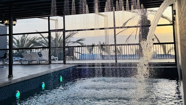 A pool area with multiple water jets, a covered seating area, and a view of palm trees and a distant landscape. - Lava villas (Ma'In)