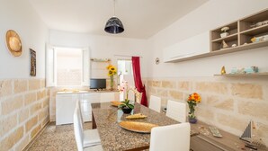 A dining area with a granite table, white chairs, a TV, and a kitchen in the background. - Holiday Home 'Residenza Della Riva Blu' with Sea View, Shared Terrace and Air Conditioning (Porto Cesareo)