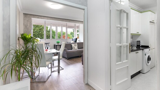 Un appartement moderne comprenant un coin repas, un canapé, une cuisine et une vue sur l’extérieur, visible à travers une porte.