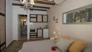 A bedroom with a bed, a bedside table with a lamp, a painting on the wall, and a view into the kitchen.