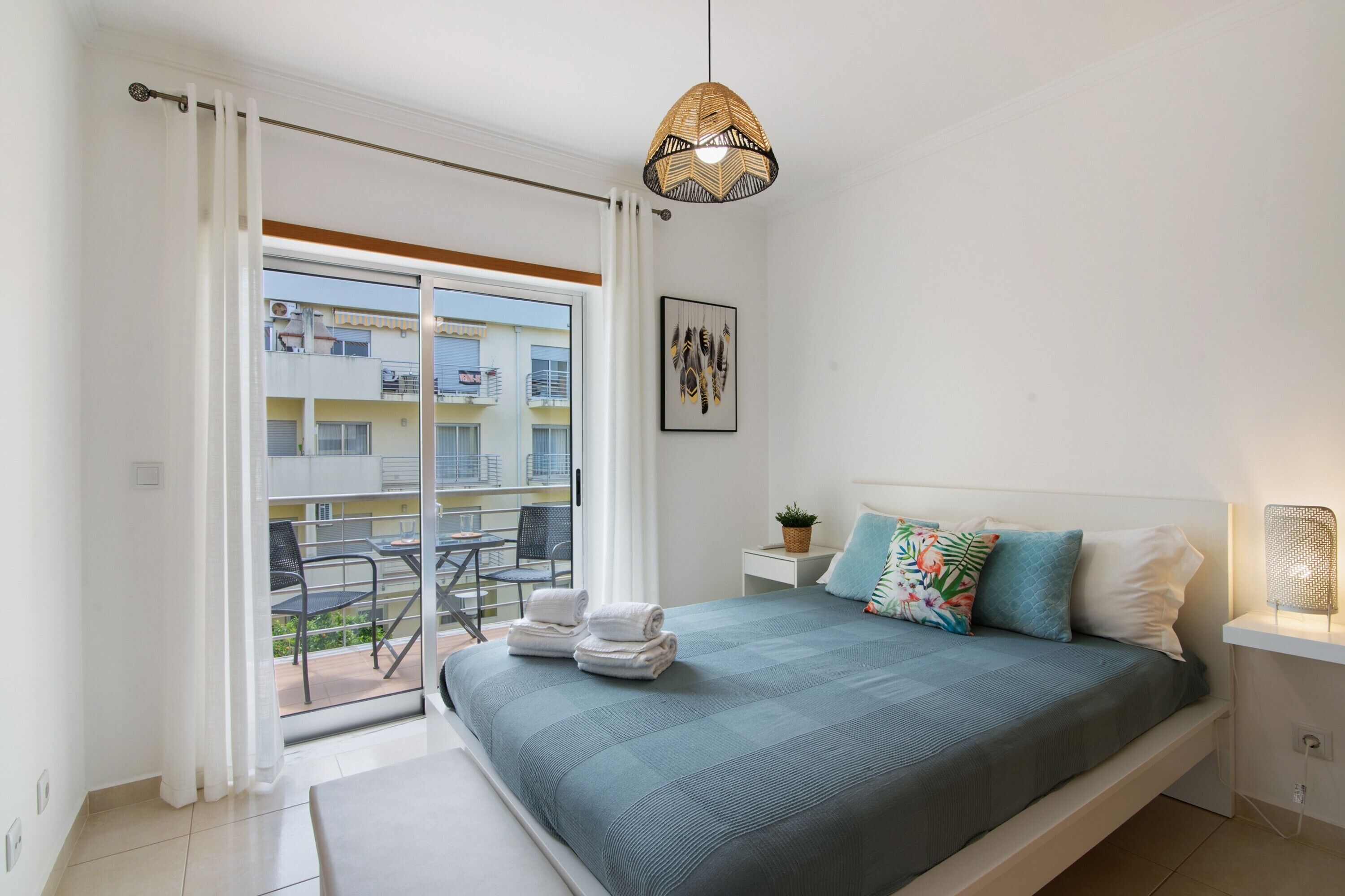 Ein Schlafzimmer mit Bett, Nachttisch, Pendelleuchte, einem Fenster mit Vorhängen und Blick auf den Balkon.