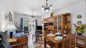 A dining area with a wooden table set for four, a television in the background, and a chandelier above. - Apartment 'Dunamar' with Private Terrace, Wi-Fi and Air Conditioning (Castro Marim)