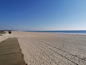Een breed zandstrand met een houten looppad dat naar een klein gebouw leidt.