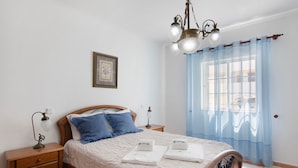 A neatly made bed with a wooden headboard, a bedside table with a lamp, a framed picture on the wall, and a window with sheer curtains. - Apartment 'Dunamar' with Private Terrace, Wi-Fi and Air Conditioning (Castro Marim)