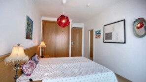 Une chambre à coucher avec un lit, deux lampes de chevet, un luminaire suspendu rouge, une porte en bois et un tableau encadré au mur.