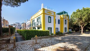 Un sendero adoquinado que conduce a un edificio amarillo y blanco con techo verde, rodeado de vegetación y otros edificios.