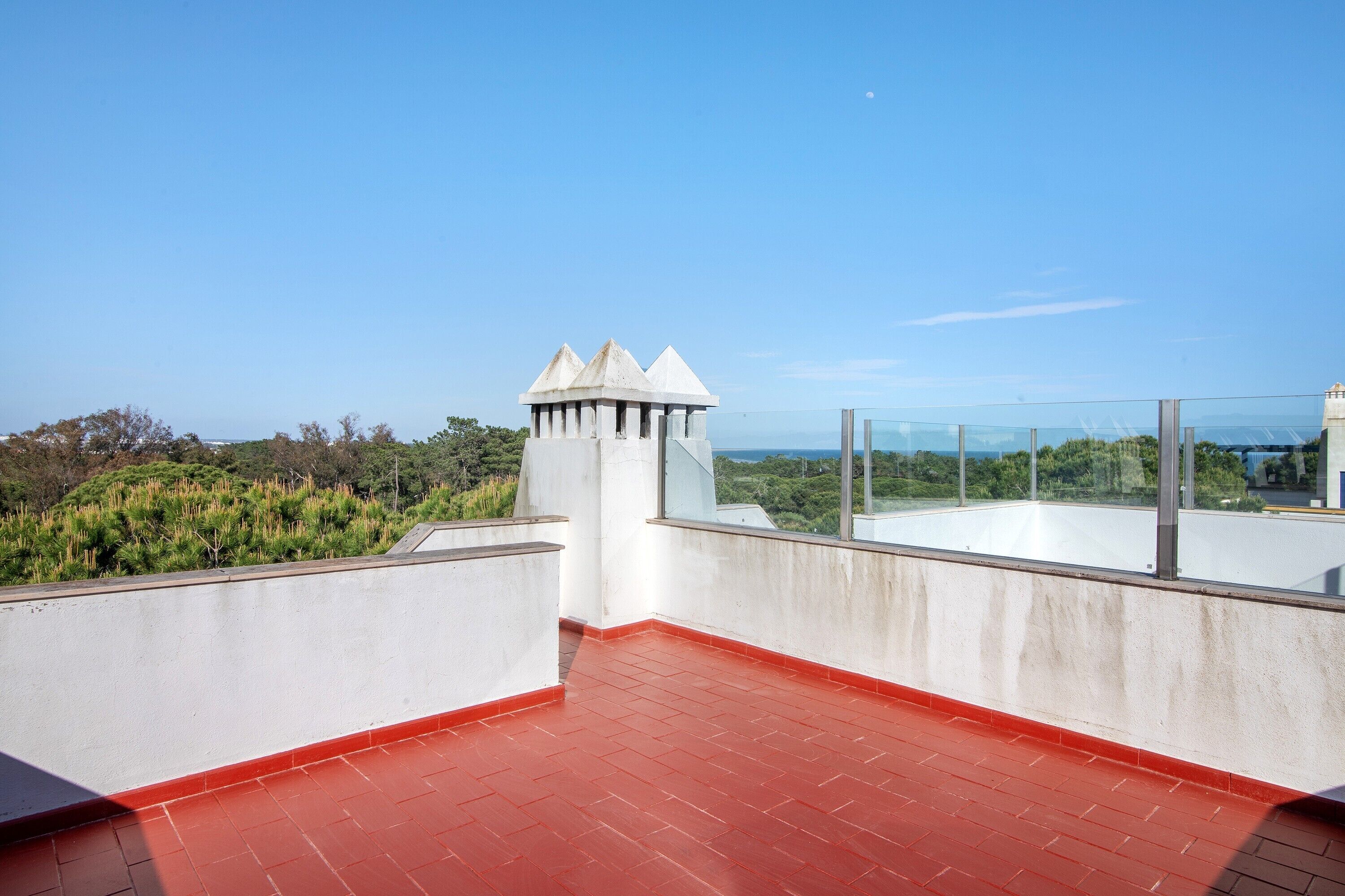 Una terraza en la azotea con piso rojo, una estructura blanca con techos puntiagudos y vista despejada a zonas verdes y edificios lejanos.