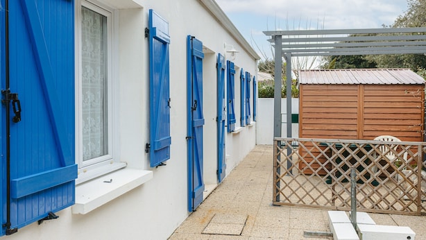 A building with white walls and blue shutters, a wooden shed, and a metal fence. - Holiday Home 'Lumineuse Près La Rémigeasse' with Private Terrace, Private Garden and Wi-Fi (Dolus-d'Oléron)