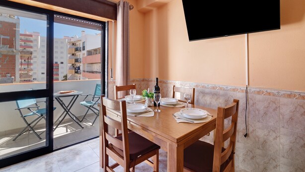 Un área de comedor con una mesa preparada para cuatro personas, un televisor montado en la pared y un balcón con vistas a edificios.
