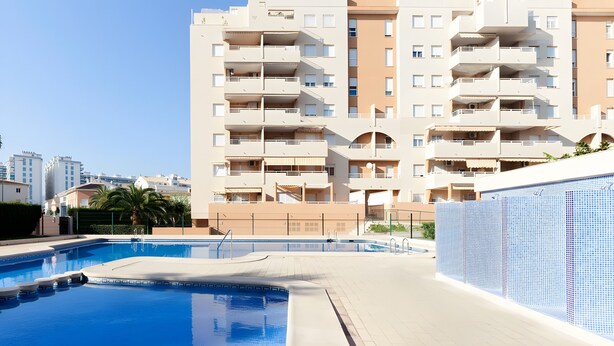 Een zwembad met betegeld zonnedek, omringd door een modern appartementencomplex met balkons en een heldere lucht.
