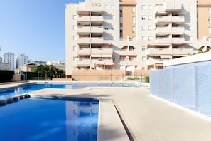 A swimming pool with a tiled deck, surrounded by a modern apartment building with balconies and a clear sky.