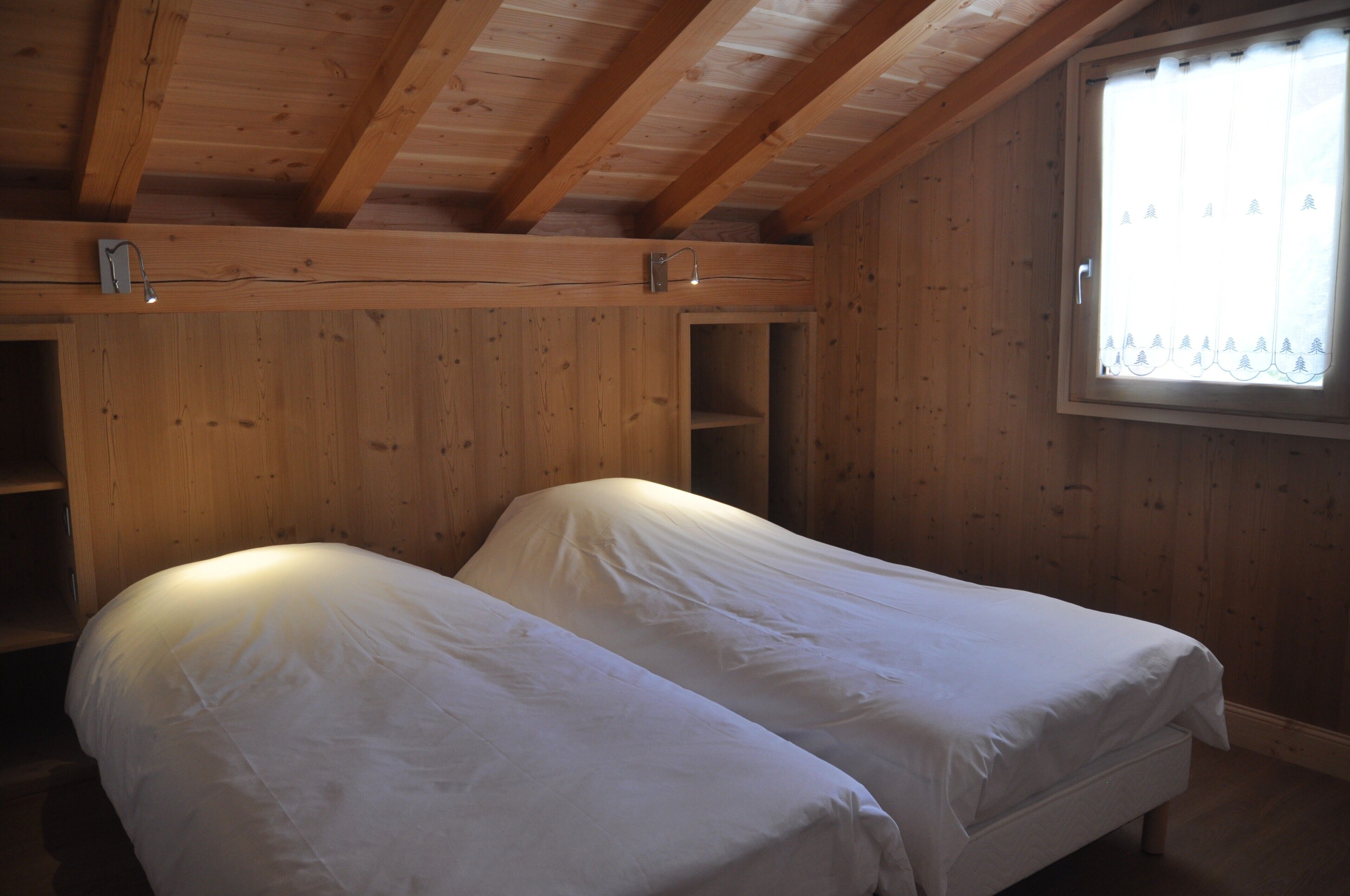 Een kamer met twee eenpersoonsbedden, houten wanden, een raam met gordijn en een schuine kap.