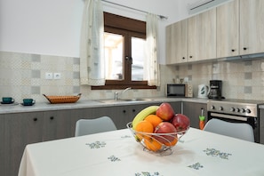 A kitchen with a dining table, a fruit bowl, and a window with curtains. - Apartment 'Iliana 1' with Mountain View, Wi-Fi and Air Conditioning (Kalymnos)