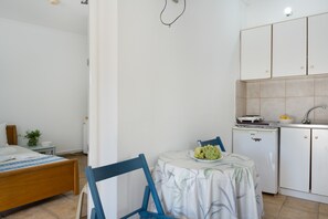 Een kleine, schone kamer met een bed, een tafel met een fruitschaal, een stoel, een kitchenette en een kleine koelkast.
