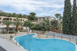 A swimming pool with a curved design, surrounded by potted plants and palm trees, with buildings in the background. - Apartment 'Studio 8' with Mountain View, Wi-Fi and Air Conditioning (Panormos Kalymnos)