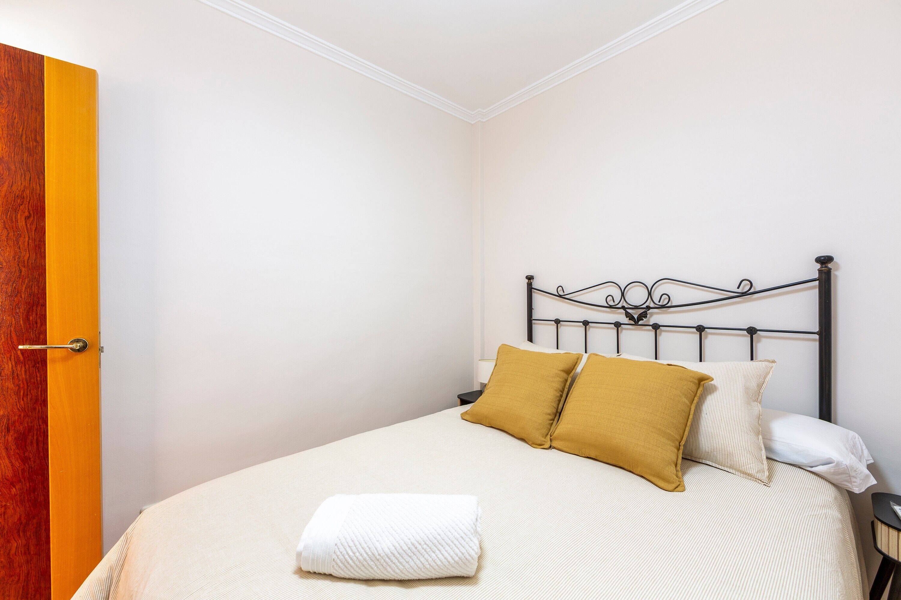 Una cama bien tendida con una manta blanca y dos almohadas, una puerta de madera y un cabecero metálico sencillo.