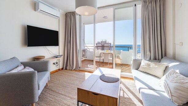 A modern living room with a flat-screen TV, grey sofas, a wooden coffee table, and a sliding glass door leading to a balcony with a view of the sea.