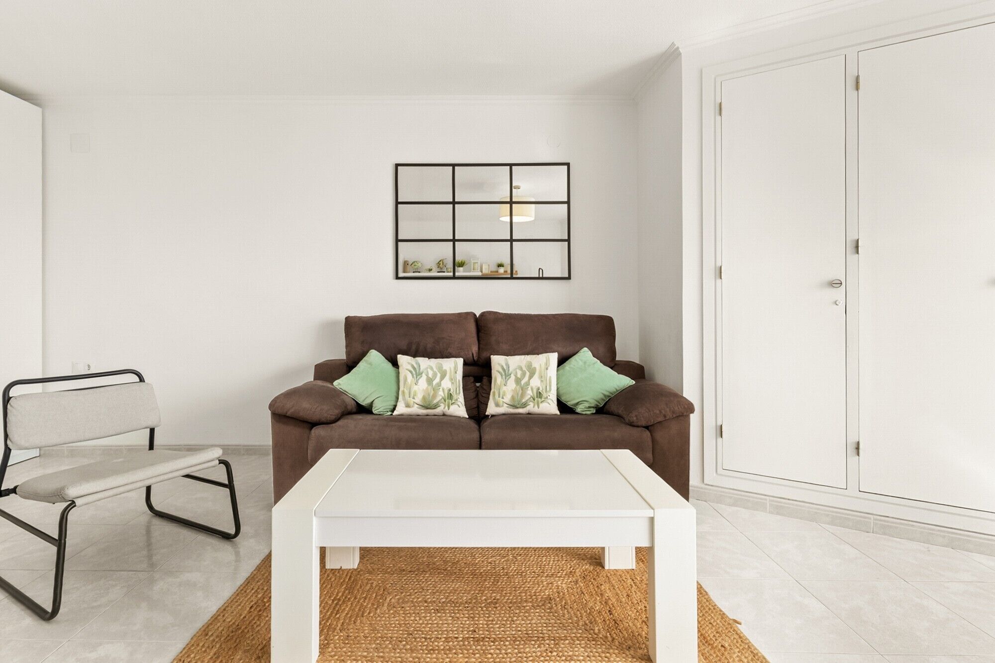 A living room with a brown sofa, a white coffee table, a woven rug, a metal-framed chair, and a white wardrobe.