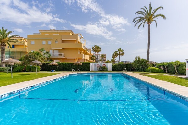 Una piscina con agua cristalina y azul, rodeada de césped cuidado, palmeras y un edificio de varios pisos con balcones.