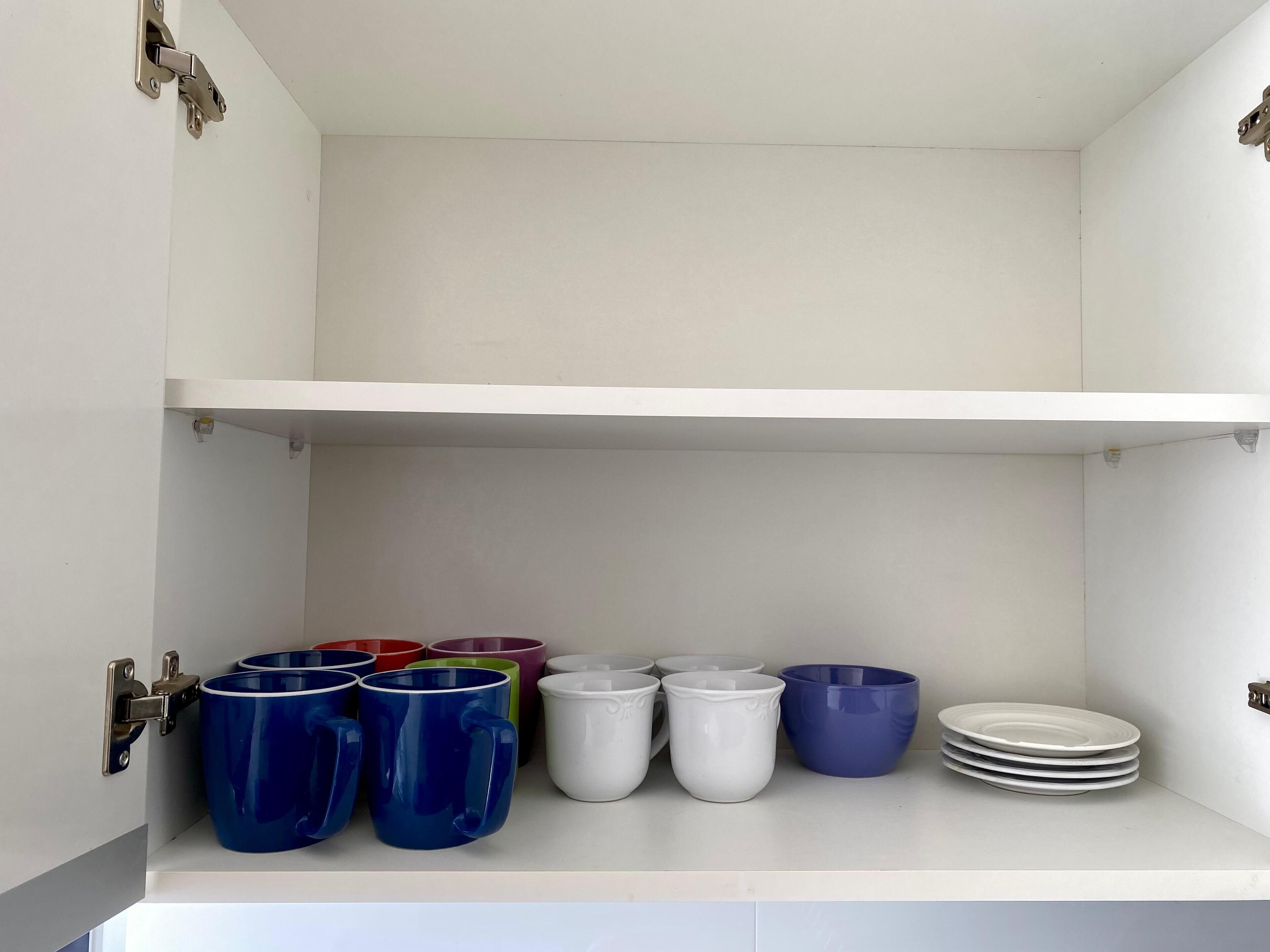 A kitchen cabinet with a white interior, containing various colored mugs and white plates.