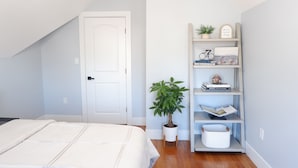 Ein Schlafzimmer mit einem Bett, einer weißen Tür, einem Bücherregal mit Büchern und Dekoration sowie einer Zimmerpflanze.