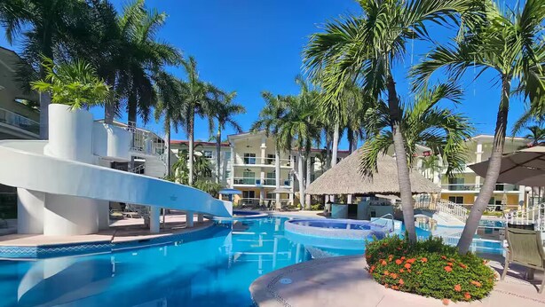 A resort pool area with multiple swimming pools, palm trees, and a thatched-roof structure.