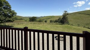 A view from a balcony with a wooden railing, overlooking a grassy landscape with rolling hills and a clear blue sky.