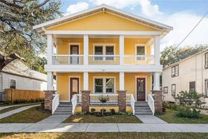 A two-story yellow house with a front porch, brick columns, and a small garden.