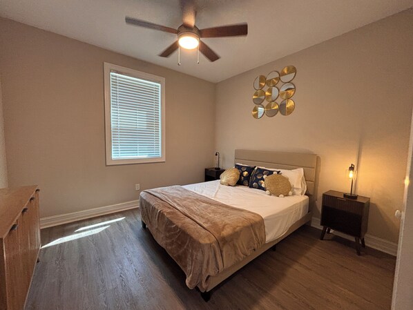 A bedroom with a bed, a ceiling fan, a nightstand, a lamp, a window with blinds, and a wall decoration.