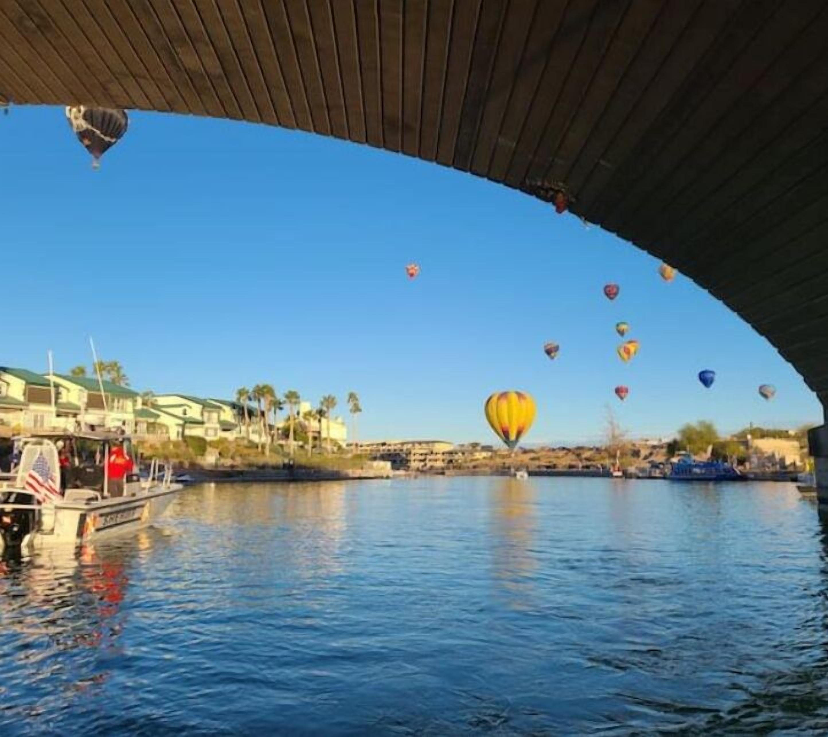 A river with hot air balloons and a boat under a bridge.