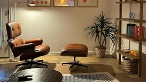 Een moderne woonkamer met een leren fauteuil, poef en een vloerlamp. Er hangen drie kunstwerken aan de muur en een boekenkast met boeken en decoratieve objecten.