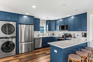 A modern kitchen with blue cabinets, stainless steel appliances, a central island, and a seating area with wicker stools.