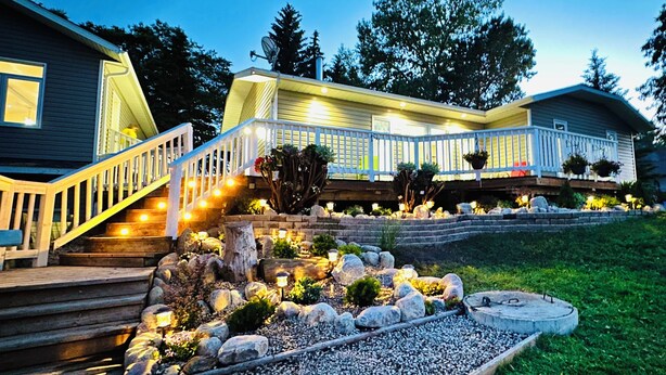Una casa de dos pisos con un porche de madera y barandal blanco. Un jardín con rocas y plantas al frente de la casa.