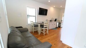 Un salon moderne avec un canapé en cuir gris, une table à manger avec des chaises, un coin cuisine avec un téléviseur fixé au-dessus du comptoir, et un plancher en bois.