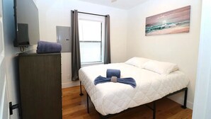 Une chambre à coucher avec un lit, une commode, un téléviseur, une fenêtre avec des rideaux et un tableau au mur.