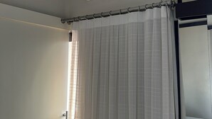 Une pièce avec un climatiseur fixé au plafond, une fenêtre avec un rideau blanc et un lit avec un matelas blanc.