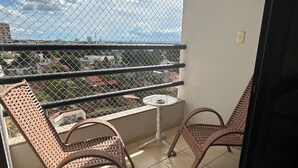 Un balcon avec des fauteuils en osier et une petite table, un sol carrelé et une vue sur la ville à travers une clôture en treillis métallique.
