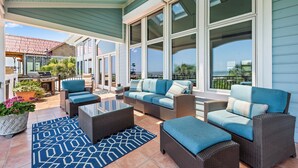 Ein überdachter Patio-Bereich mit Korbmöbeln, einem Glastisch und einem blauen Teppich mit Muster.