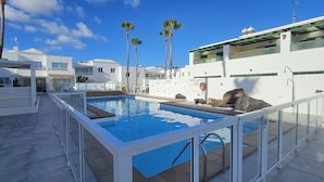 A swimming pool with a white deck, surrounded by white buildings with balconies, palm trees, and a clear blue sky.
