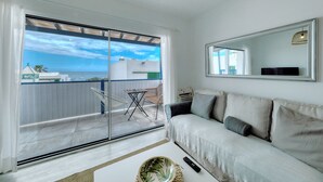A room with a sliding glass door leading to a balcony, a sofa, a mirror, and a remote control on the floor.
