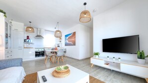A modern living room with a flat-screen TV, a dining area with a table and chairs, and a kitchen in the background.