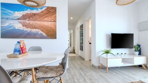 A modern living room with a white sofa, a dining table with chairs, a flat-screen TV, and a large beach painting.
