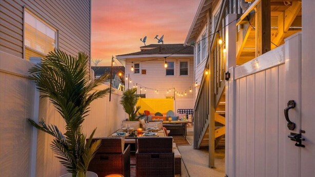 A patio with a dining table set for two, surrounded by chairs, a potted plant, and string lights.