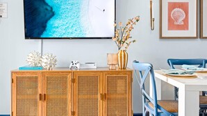 A room with a television displaying a beach scene, a wooden cabinet with decorative items, a vase with flowers, a blue chair, and a framed picture on the wall.