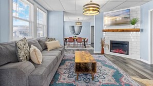 A modern living room with a gray sofa, a wooden coffee table, a fireplace, and a dining area in the background.