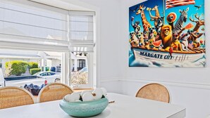 A dining area with a white table, wicker chairs, and a bowl of white fruits. A colorful mural of animals and the text "MARGATE CITY" is on the wall. - 3BR Home - Close to Beach - Backyard Oasis (Margate City)