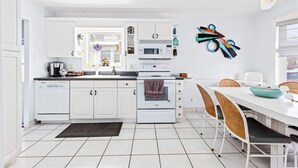 A modern kitchen with white cabinetry, a dining area with wicker chairs, and a colorful abstract wall art piece. - 3BR Home - Close to Beach - Backyard Oasis (Margate City)